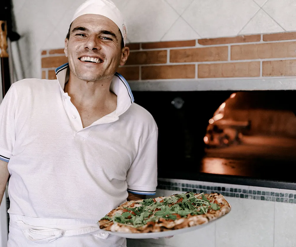 Il pizzaiolo sorride con una pizza davanti al forno a legna